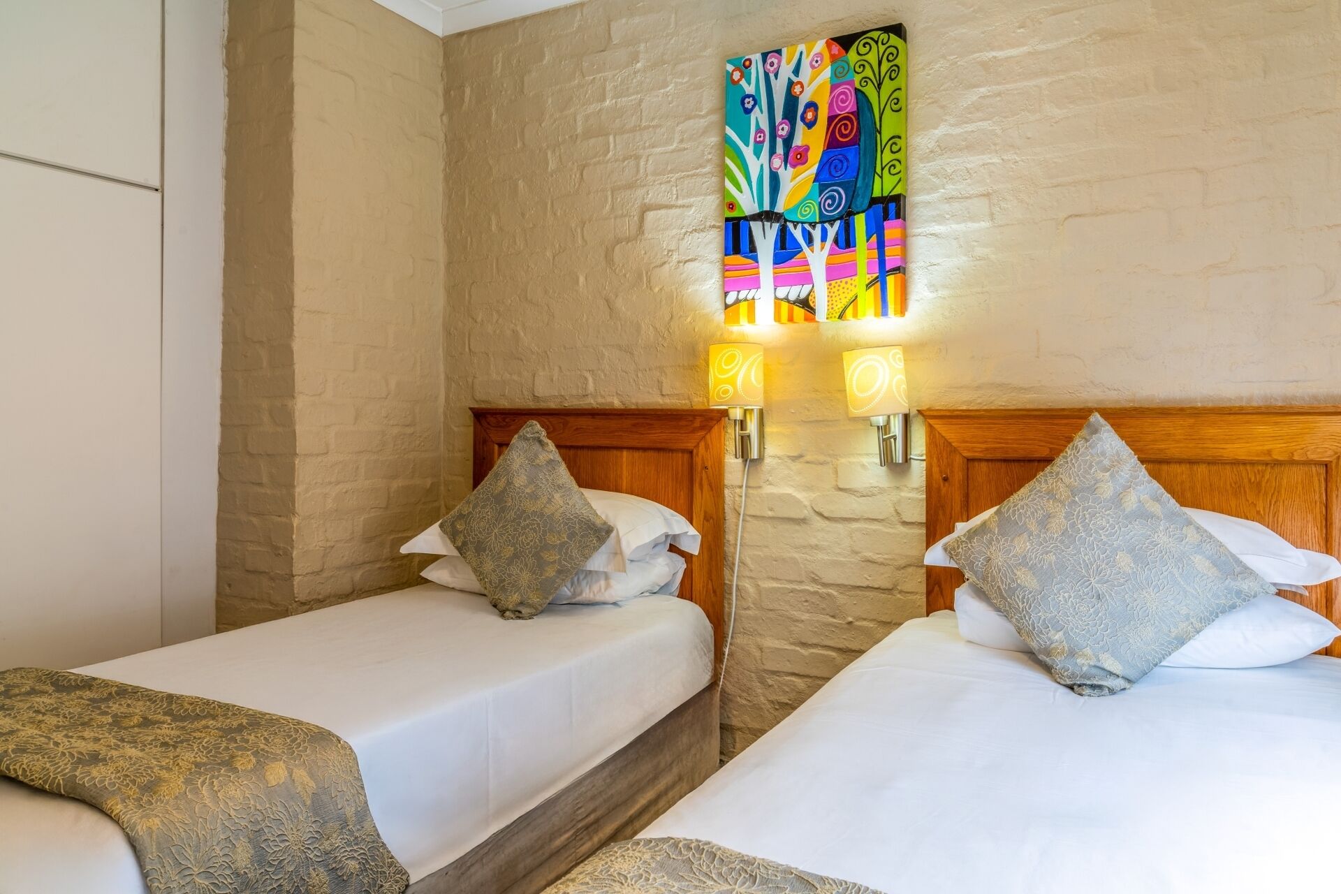 A hotel room with two beds, each with a wooden headboard, white bedding, and patterned pillows. There are wall sconces with decorative lamps, and a colorful abstract painting on the wall.