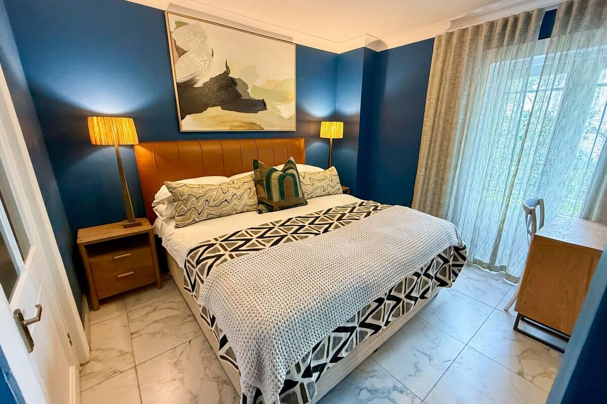 A bedroom with a bed, nightstands, a headboard, a painting, a lamp, and a window with curtains.