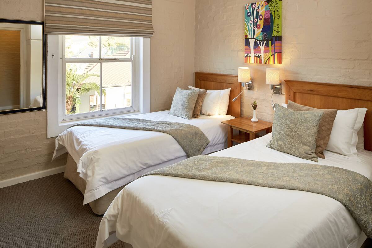 A hotel room with two beds, a window with blinds, a nightstand with lamps, and a colorful wall art piece.