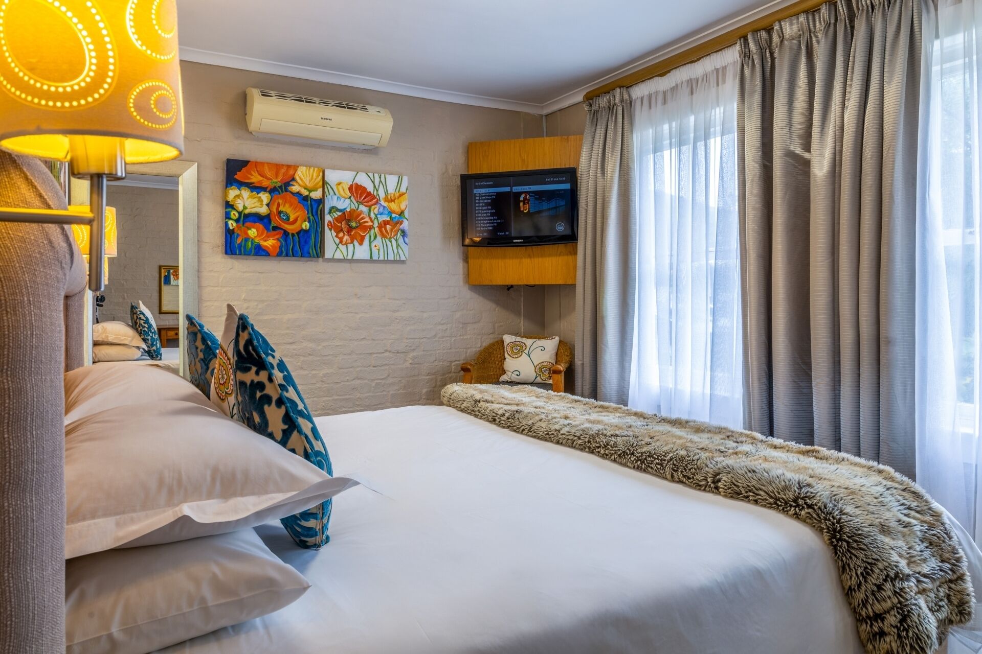 Habitación de hotel con una cama grande, un televisor de pantalla plana, aire acondicionado de pared y una ventana con cortinas traslúcidas.