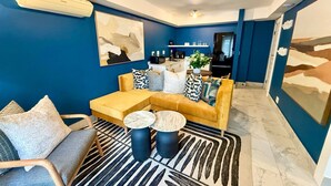Ett modernt vardagsrum med en soffa, ett soffbord och stolar, ett köksområde i bakgrunden och en stor, abstrakt målning på väggen.
