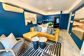 A modern living room with a sofa, coffee table, and chairs, a kitchen area in the background, and a large abstract painting on the wall. - First Group Hastings Hall (Cape Town)