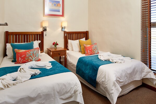 Two single beds with wooden headboards, white bedding, and colorful pillows. There are two wall-mounted lamps and a small table with a vase of flowers. - First Group Port Owen Marina (Bergrivier)