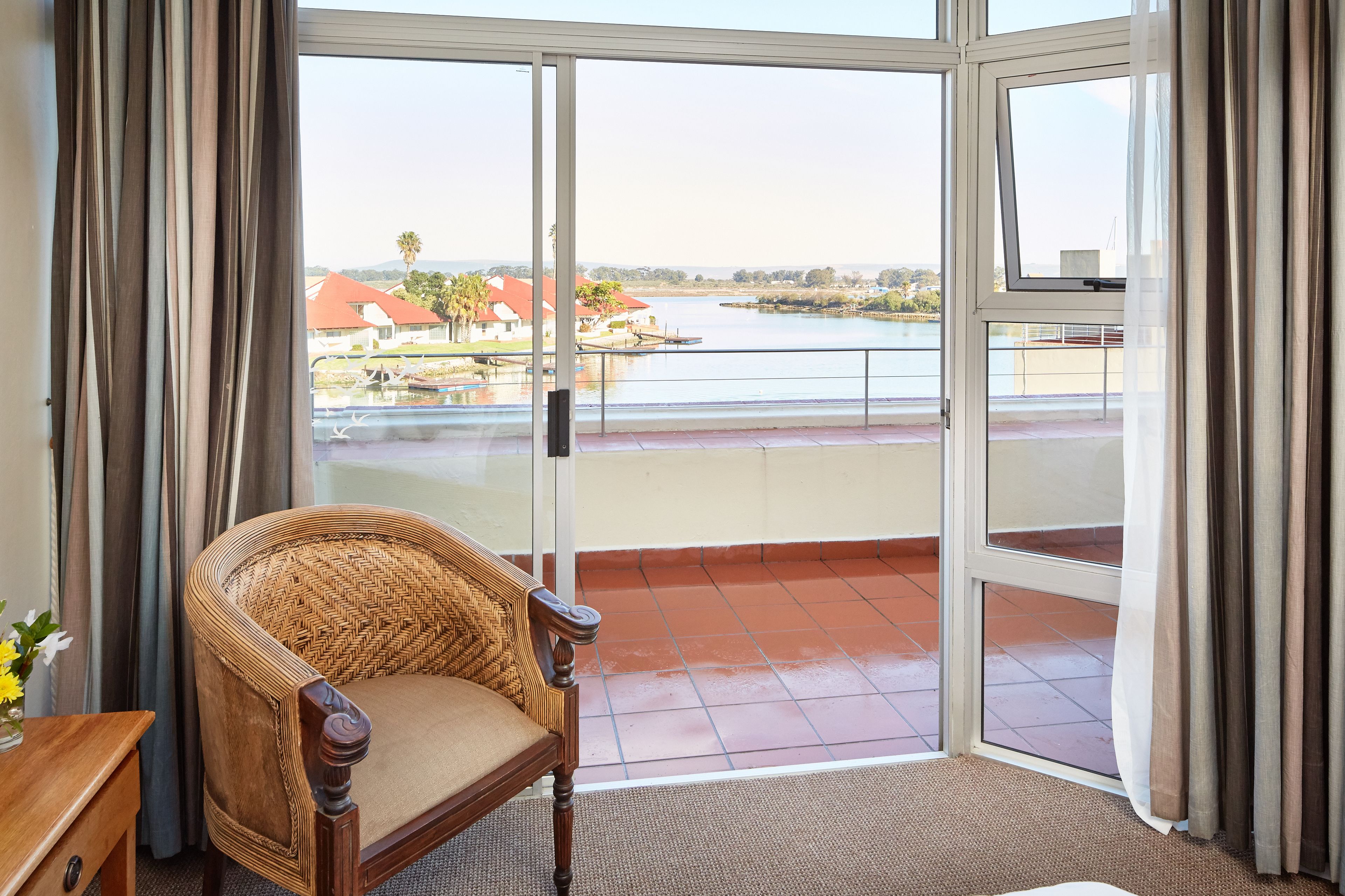 Ein Zimmer mit einem Korbstuhl, einem kleinen Tisch mit einer Vase und Blick auf einen See sowie Gebäude durch eine Glastür.