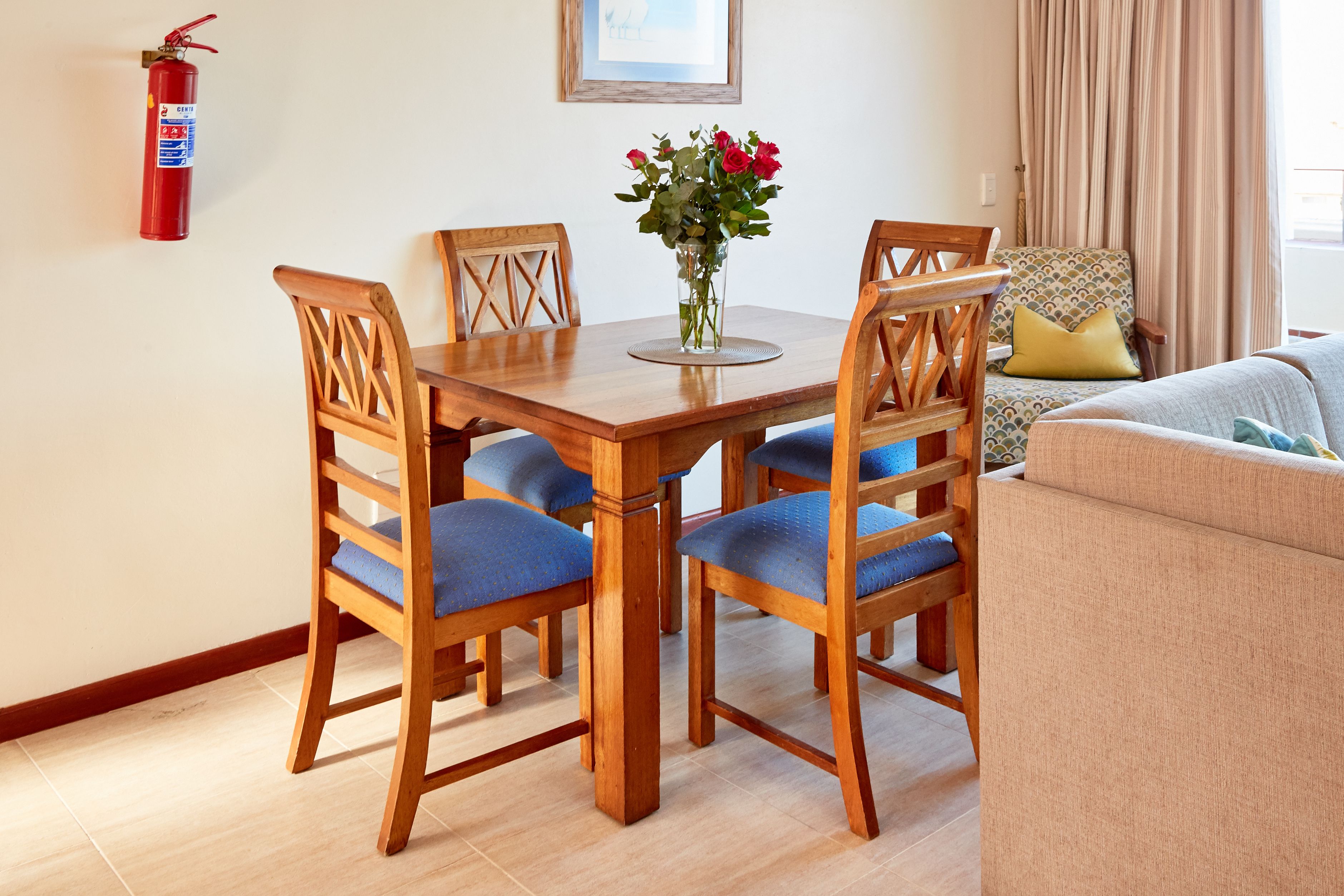 A dining area with a wooden table and chairs, a vase with flowers, a fire extinguisher, and a sofa in the background.