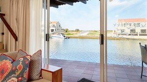 A modern living room with a sofa and cushions, a wooden staircase, and a view of a canal with boats and buildings. - First Group Port Owen Marina (Bergrivier)