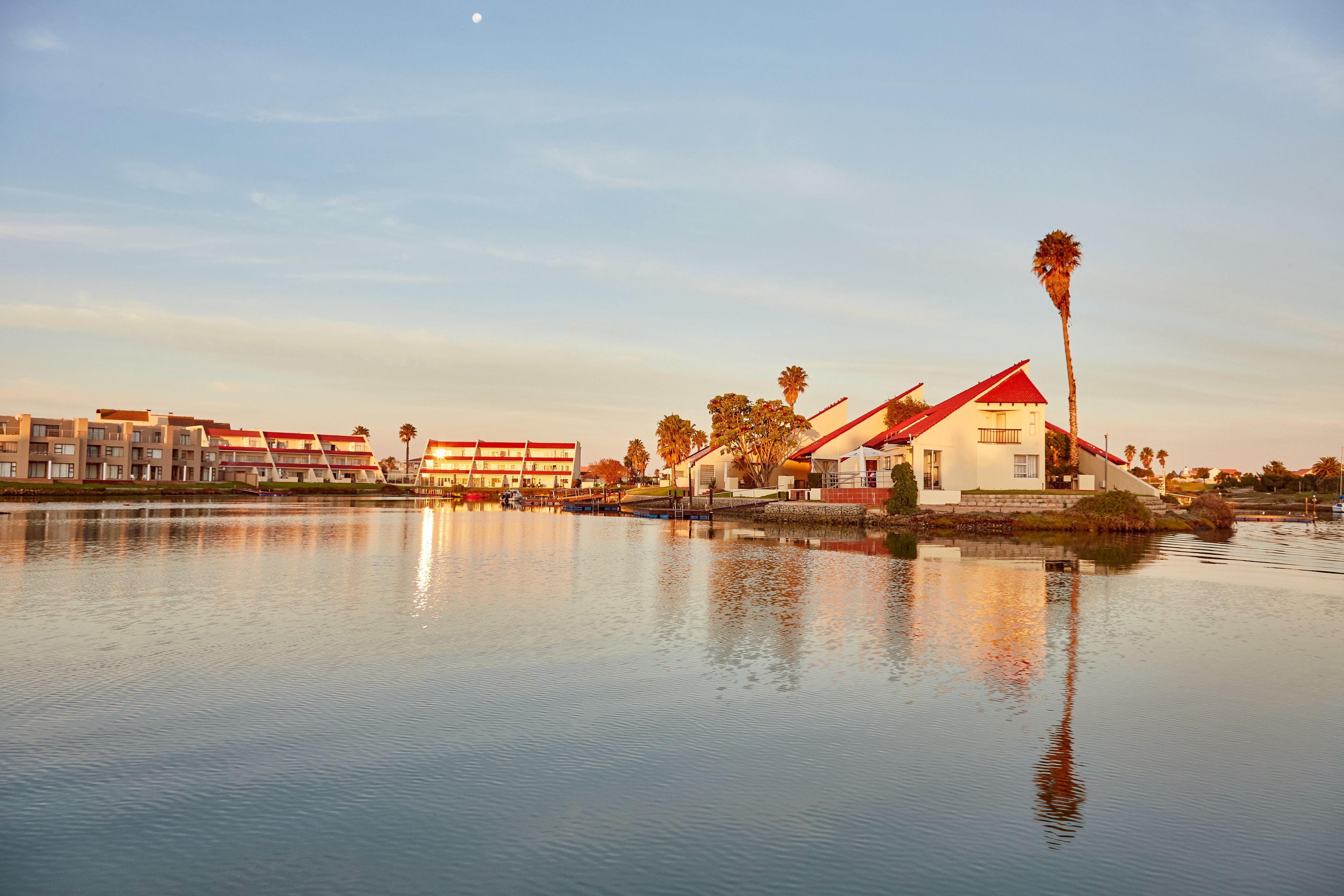 Hotel à beira-mar com um edifício de telhado vermelho, um lago tranquilo refletindo as construções e um céu claro.