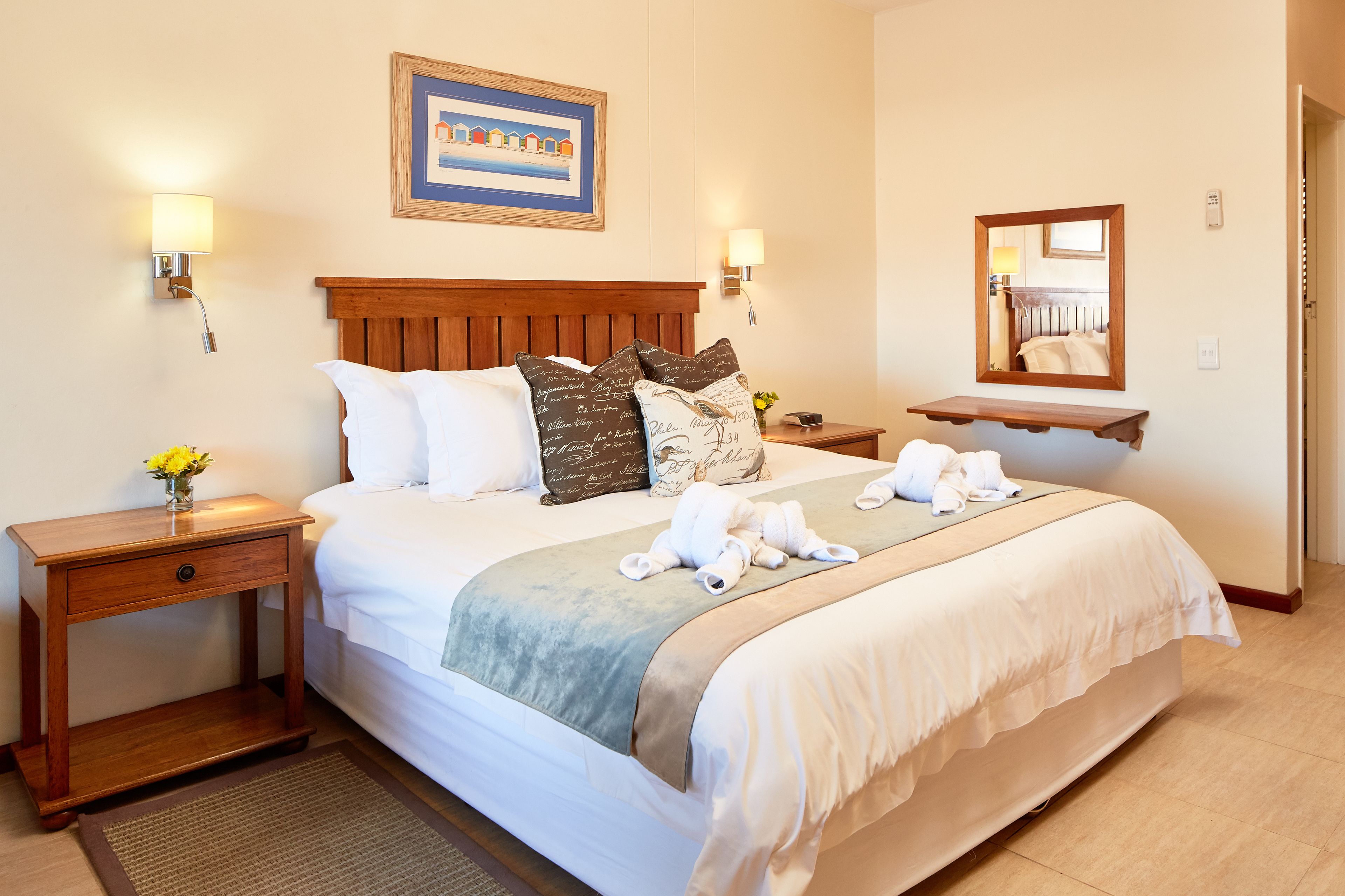 A neatly made bed with a wooden headboard, a bedside table with a lamp, a mirror, and a framed picture on the wall.
