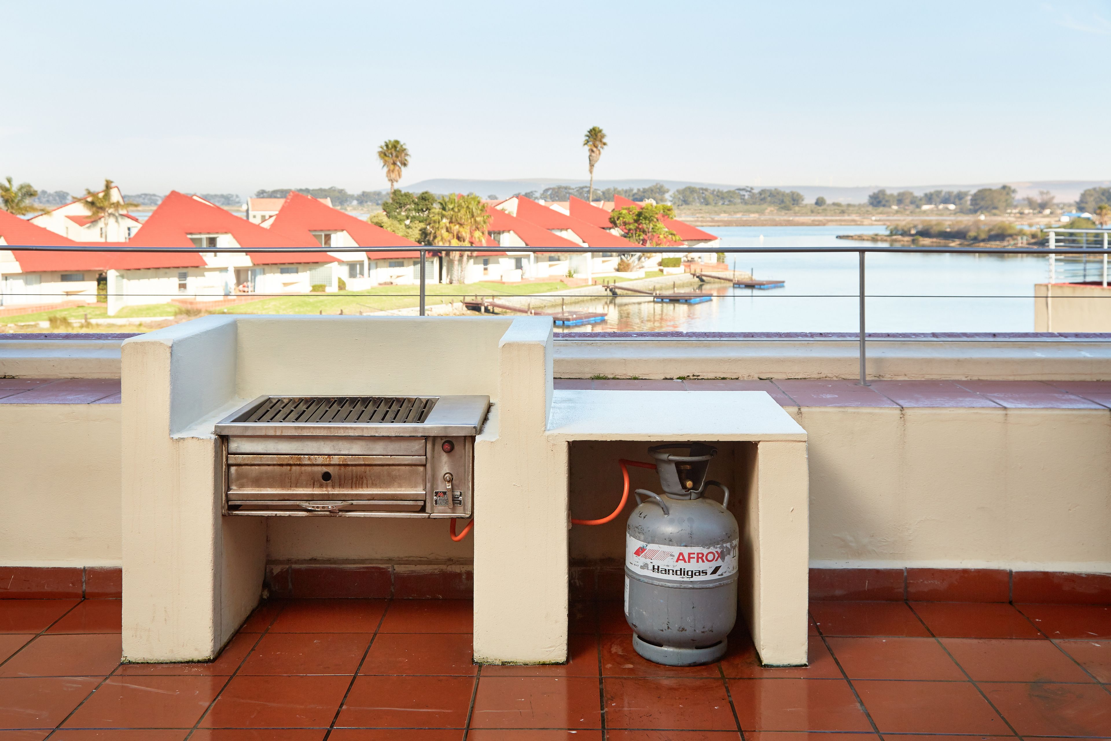 Un barbecue et une bouteille de propane sur une terrasse offrant une vue sur un lac et, au loin, sur des bâtiments aux toits rouges.