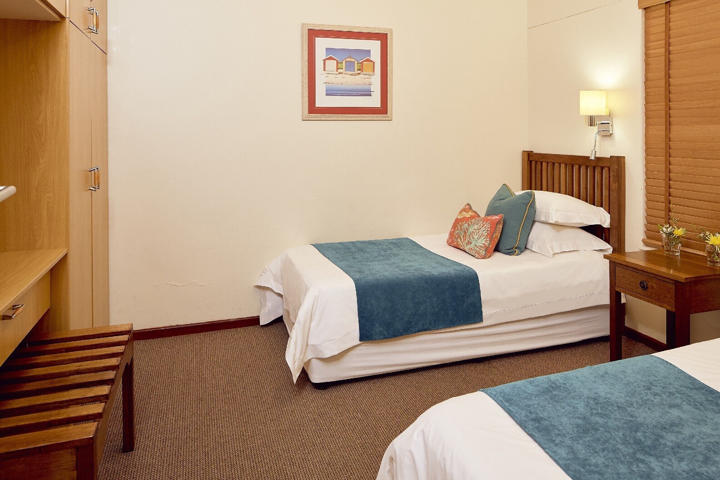 A hotel room with two beds, a wooden bench, a bedside table with a lamp, and a framed picture on the wall.