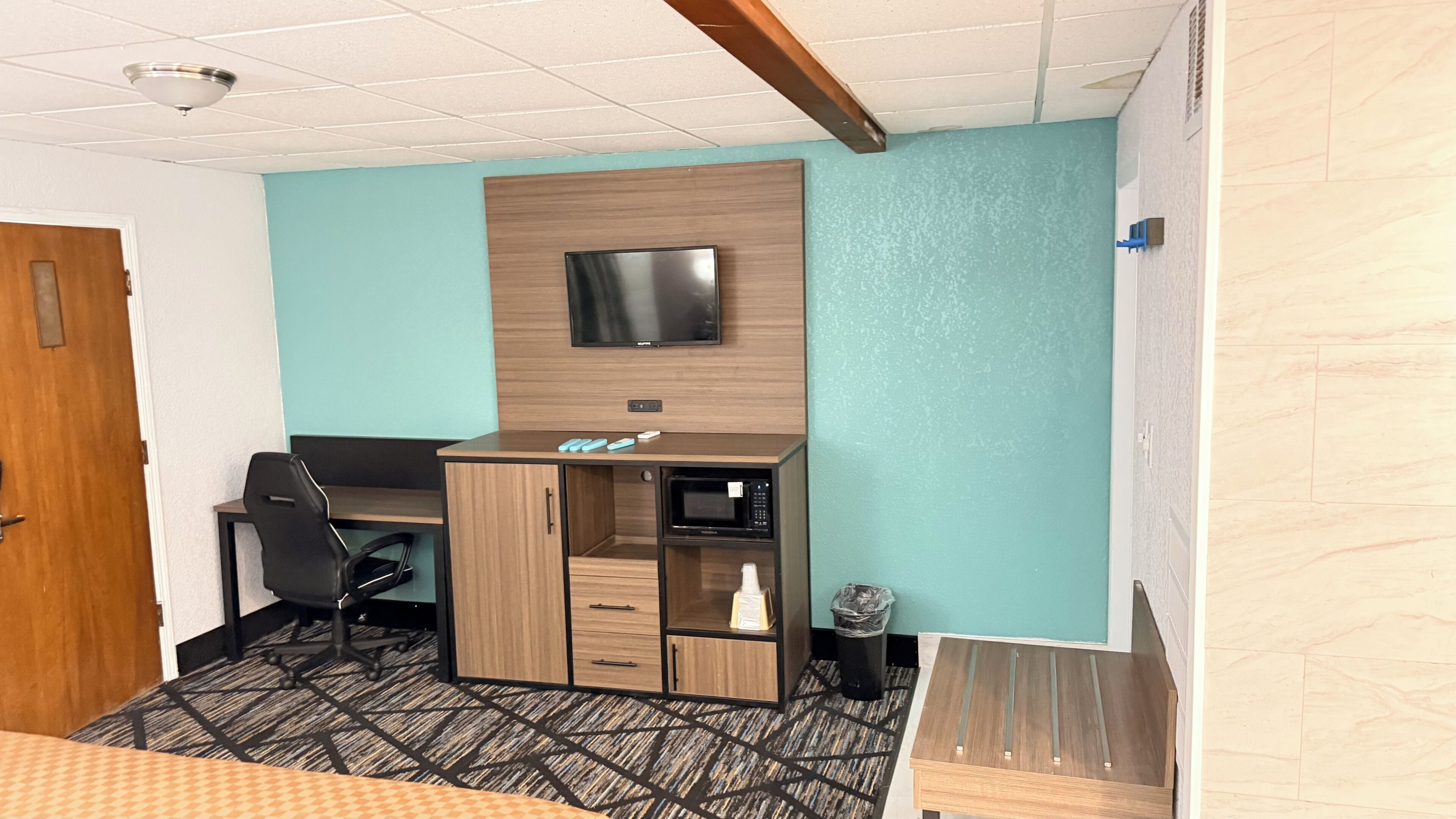 Quarto de hotel com escrivaninha de madeira, televisão, micro-ondas e lixeira.