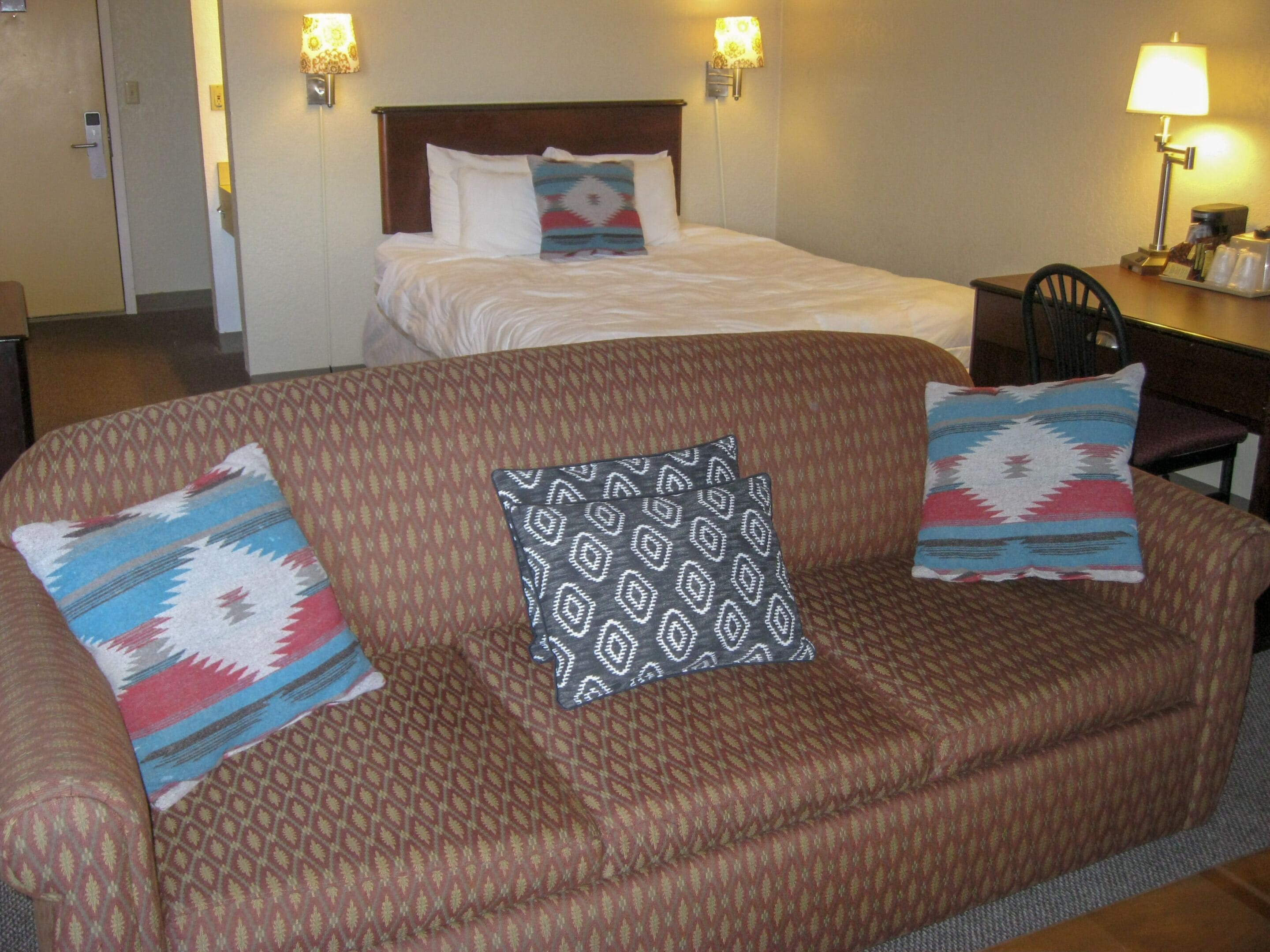 A hotel room with a large brown sofa, two patterned pillows, a bed with white bedding, and two wall-mounted lamps.