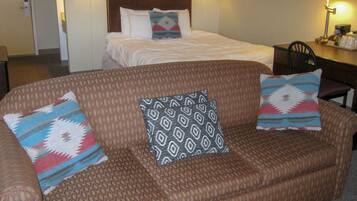 A hotel room with a large brown sofa, two patterned pillows, a bed with white bedding, and two wall-mounted lamps.