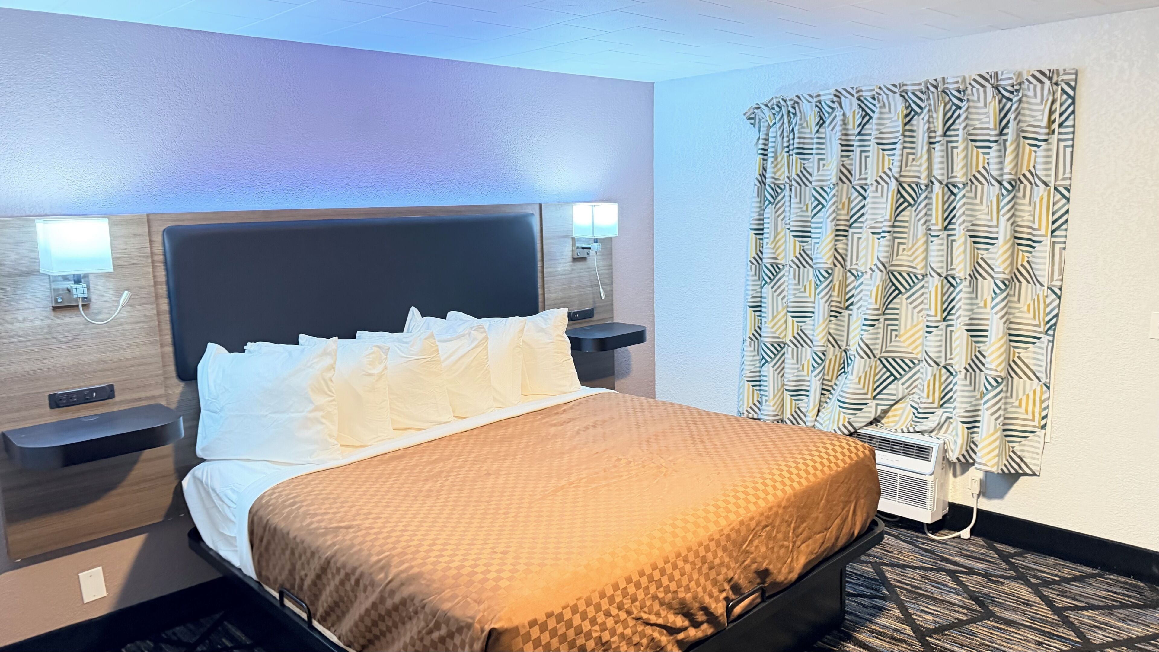 A hotel room with a bed, bedside tables, a wall-mounted lamp, a window with patterned curtains, and a wall-mounted air conditioning unit.