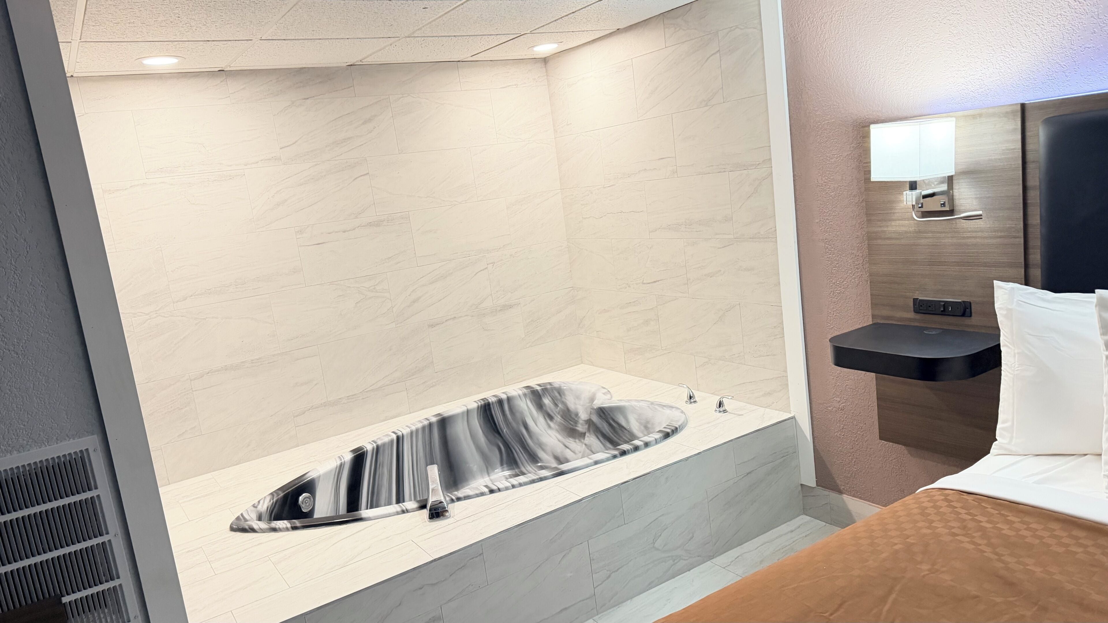 A modern bathroom with a marble wall, a freestanding bathtub, a bedside table with a lamp, and a bed with a brown headboard.