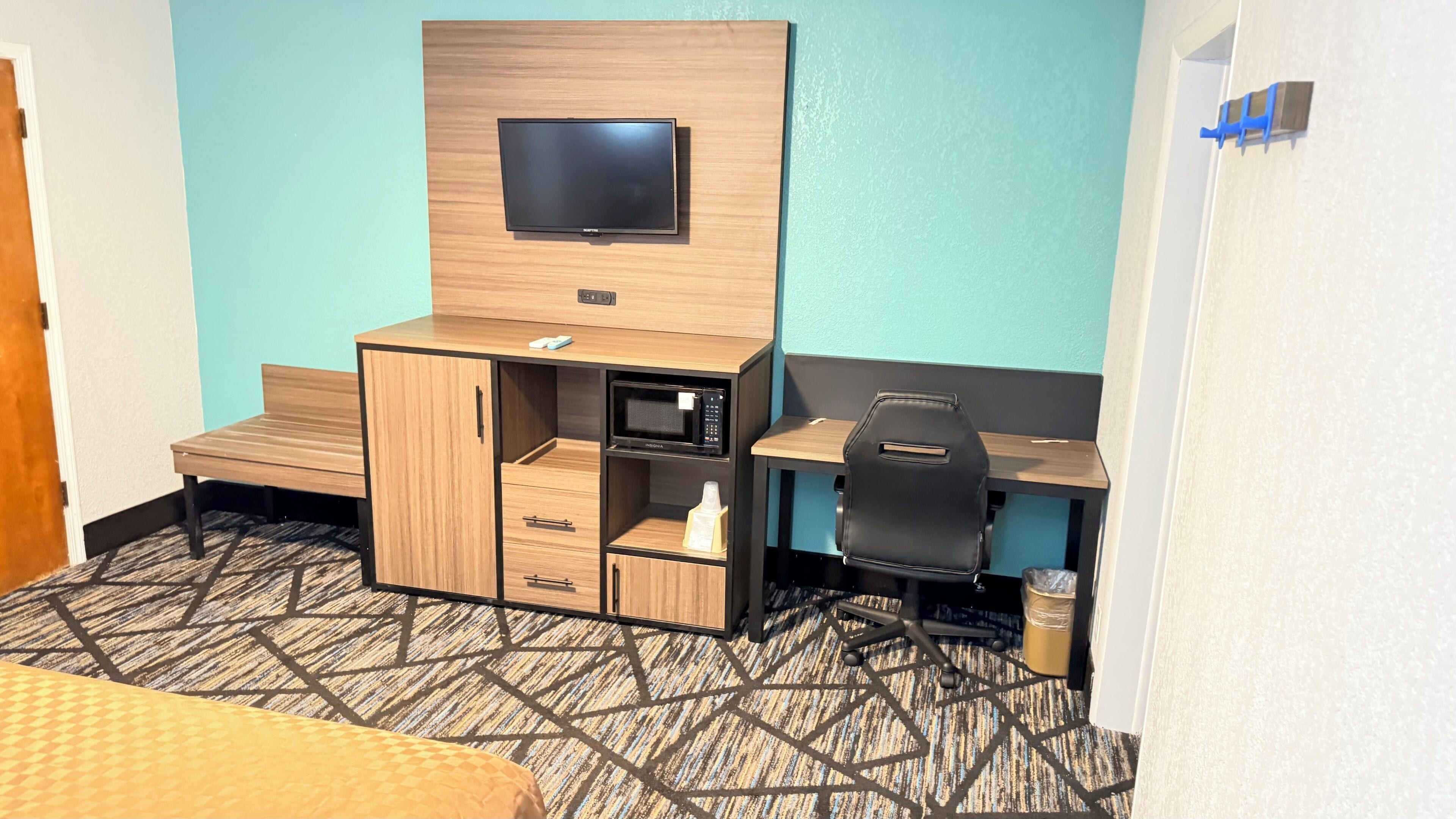 A hotel room with a bed, a desk with a flat-screen TV, a microwave, a chair, and a trash can.
