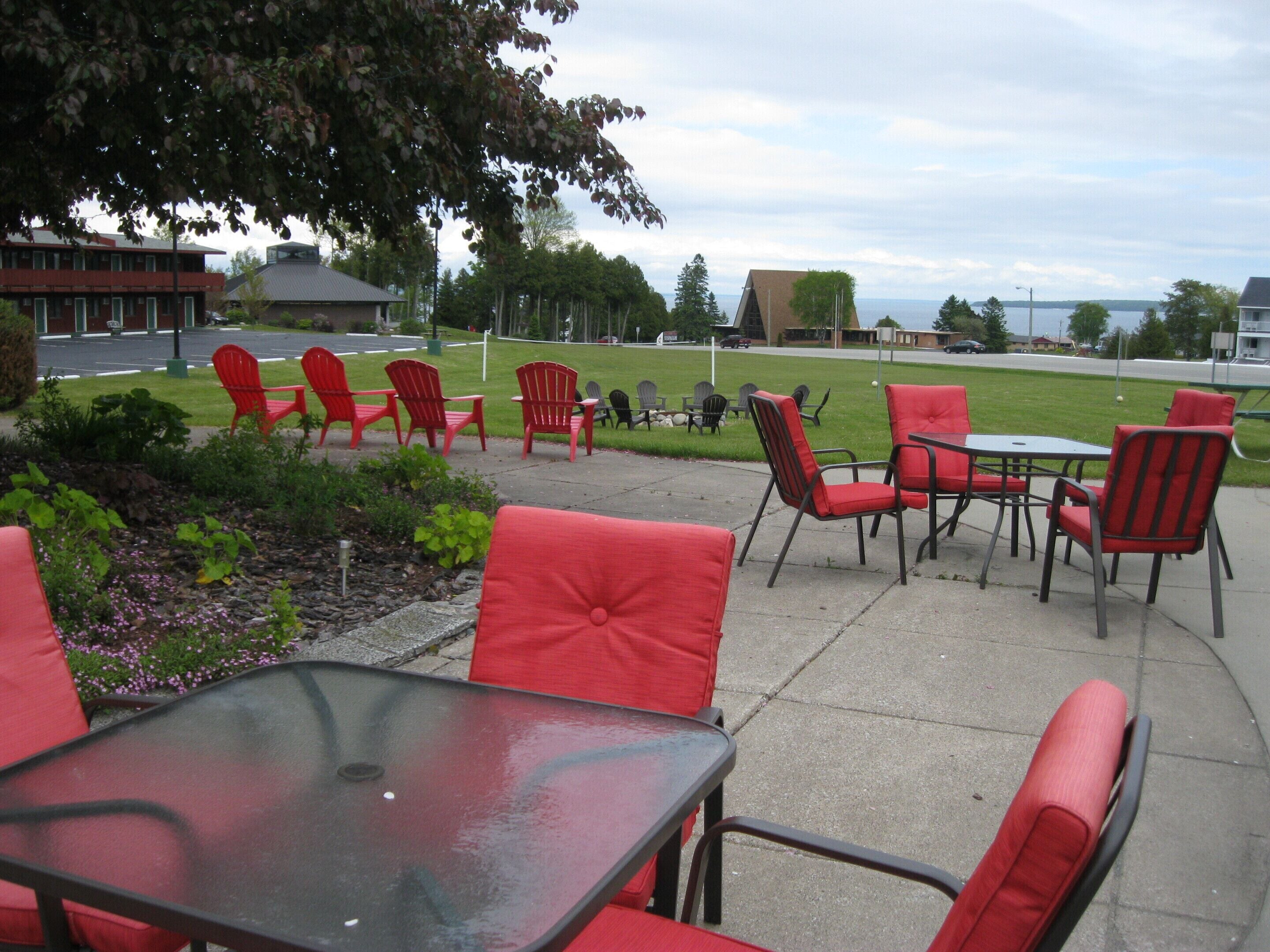 Un espace extérieur aménagé avec des chaises rouges et une table en verre, offrant une vue sur un champ verdoyant et une étendue d’eau.