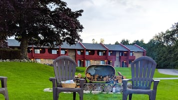A fire pit with logs, surrounded by rocks, and two Adirondack chairs facing a building with a red roof.