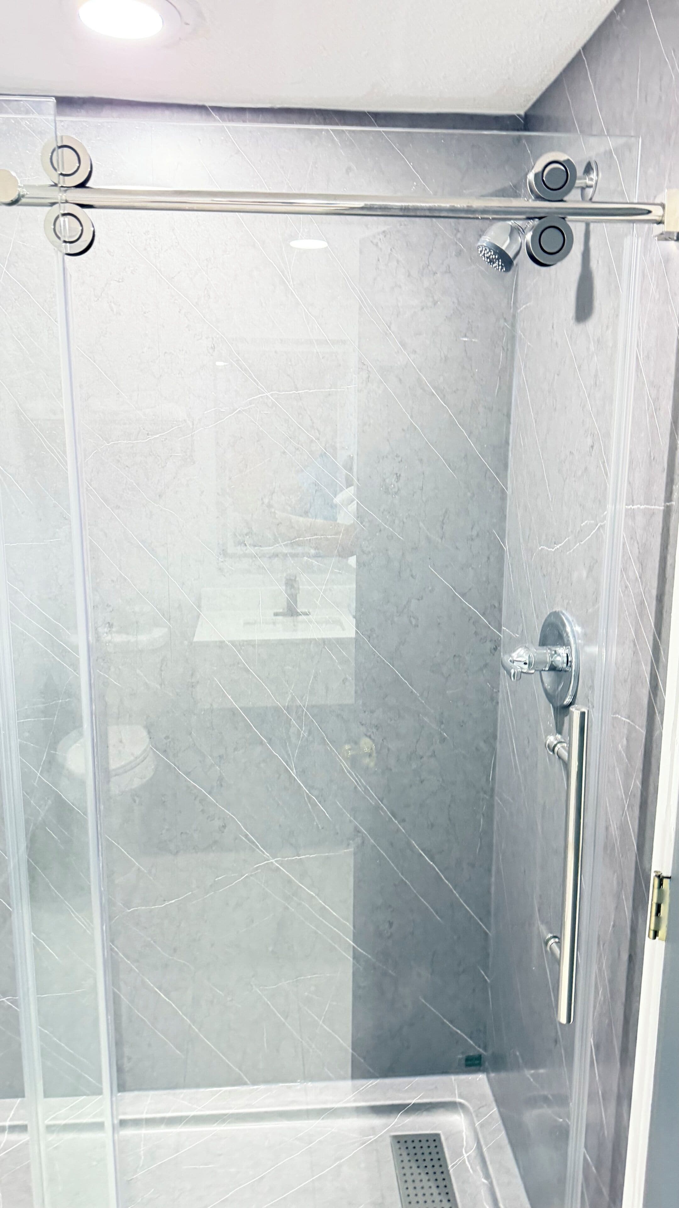 A glass-enclosed shower stall with a chrome shower head and handle, a visible drain, and a tiled wall.