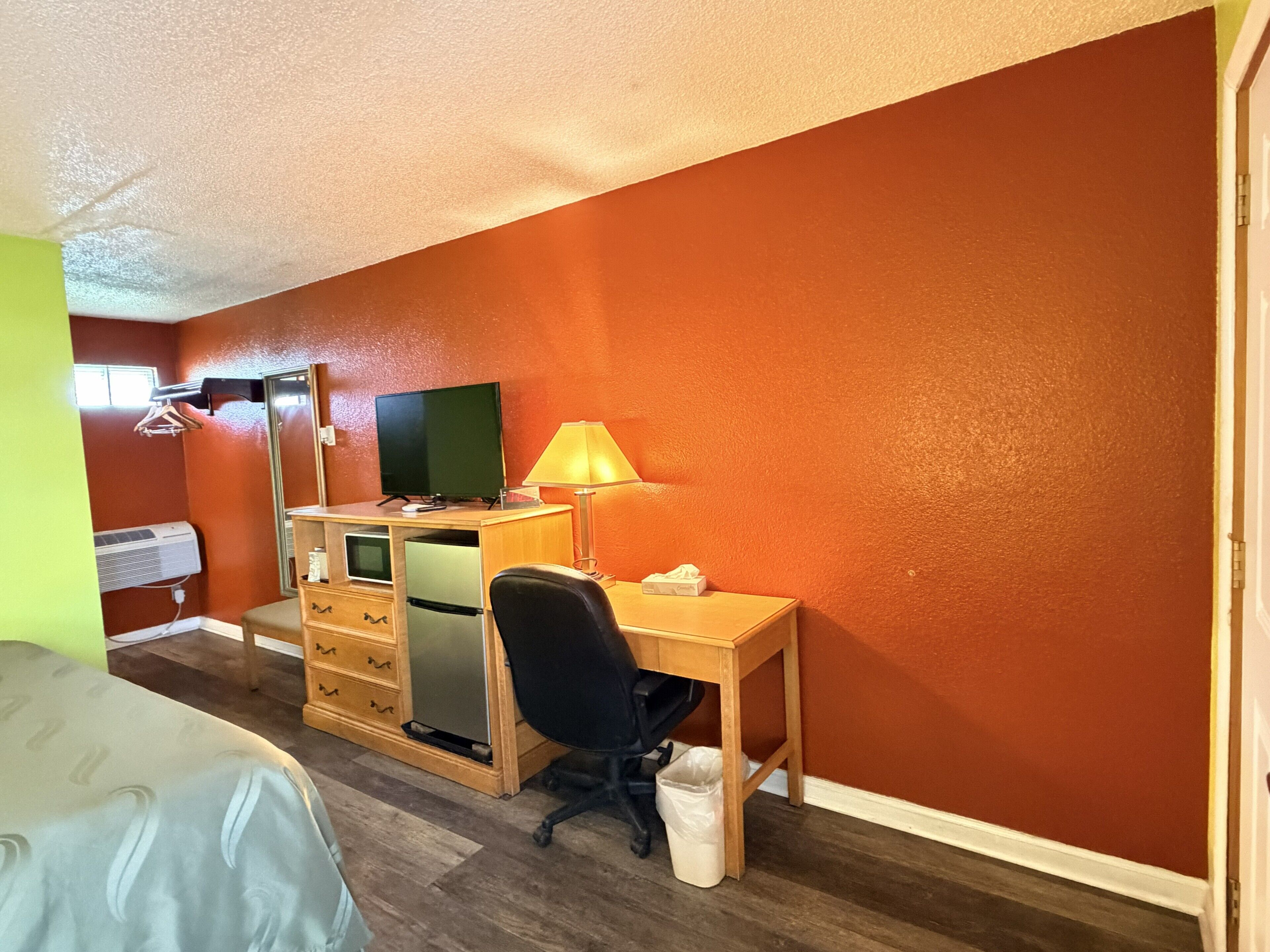 A compact hotel room with a single bed, a desk with a chair, a television, a lamp, and a small trash bin.
