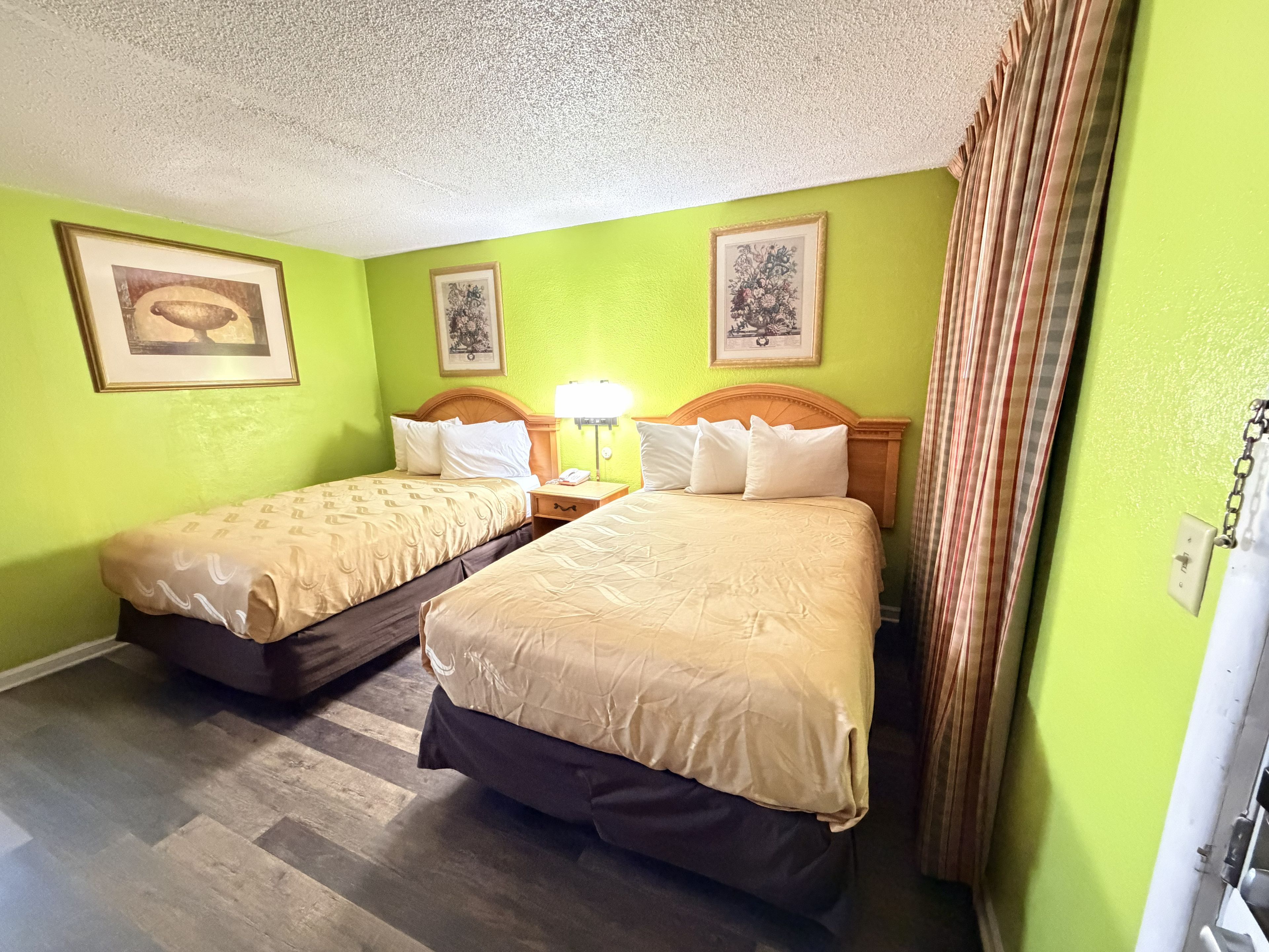 A hotel room with two beds, a wooden headboard, a nightstand with a lamp, and a green wall.