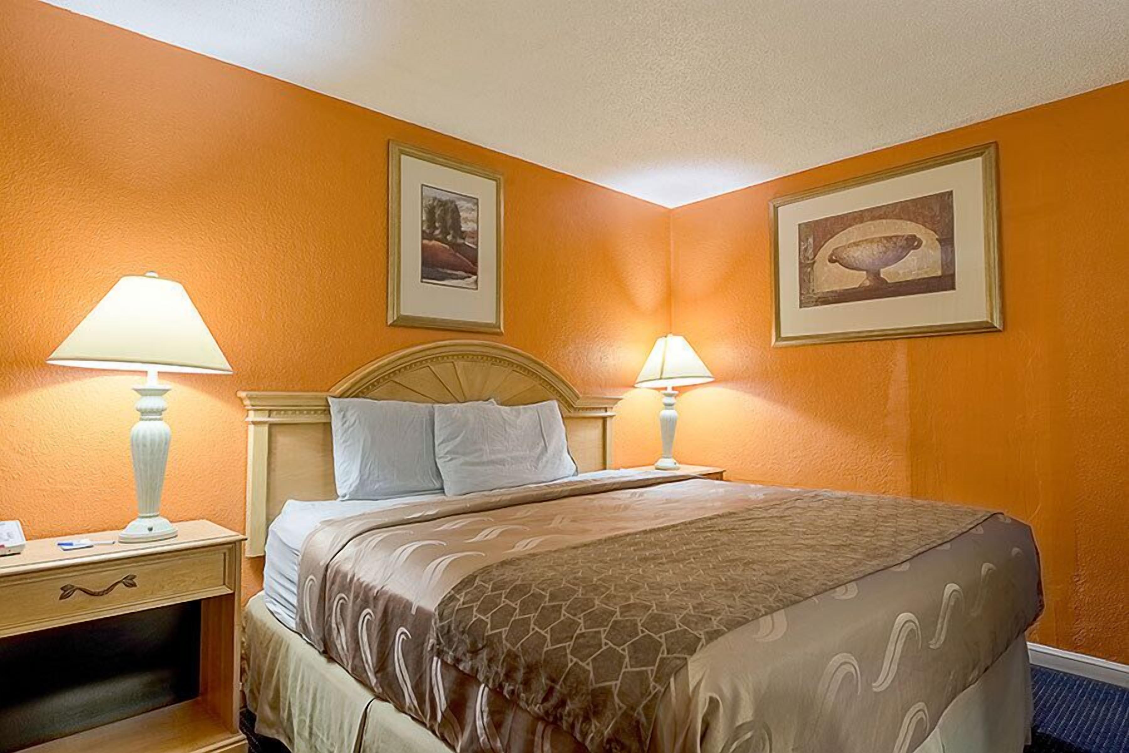 Une chambre d’hôtel avec un lit, deux lampes de chevet, une table de chevet et deux tableaux encadrés accrochés aux murs.