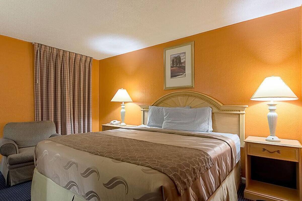 Une chambre d’hôtel comprenant un lit, deux tables de chevet avec des lampes, une chaise et une grande fenêtre avec des rideaux.