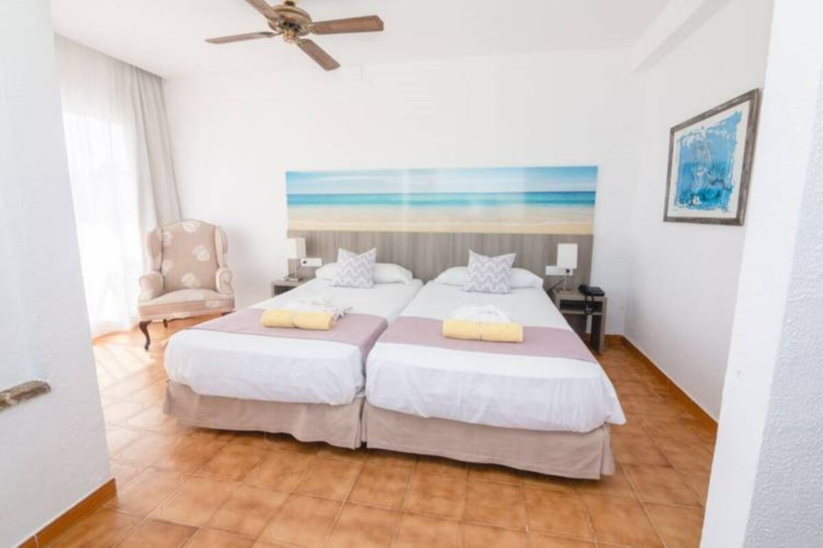 Habitación de hotel con dos camas, un ventilador de techo, un cuadro con motivos playeros, una silla y un marco con una fotografía en la pared.