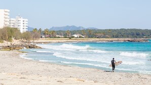 A beach with a person holding a surfboard, a rocky shoreline, and a building in the background. - Hotel Club S'illot (Manacor)