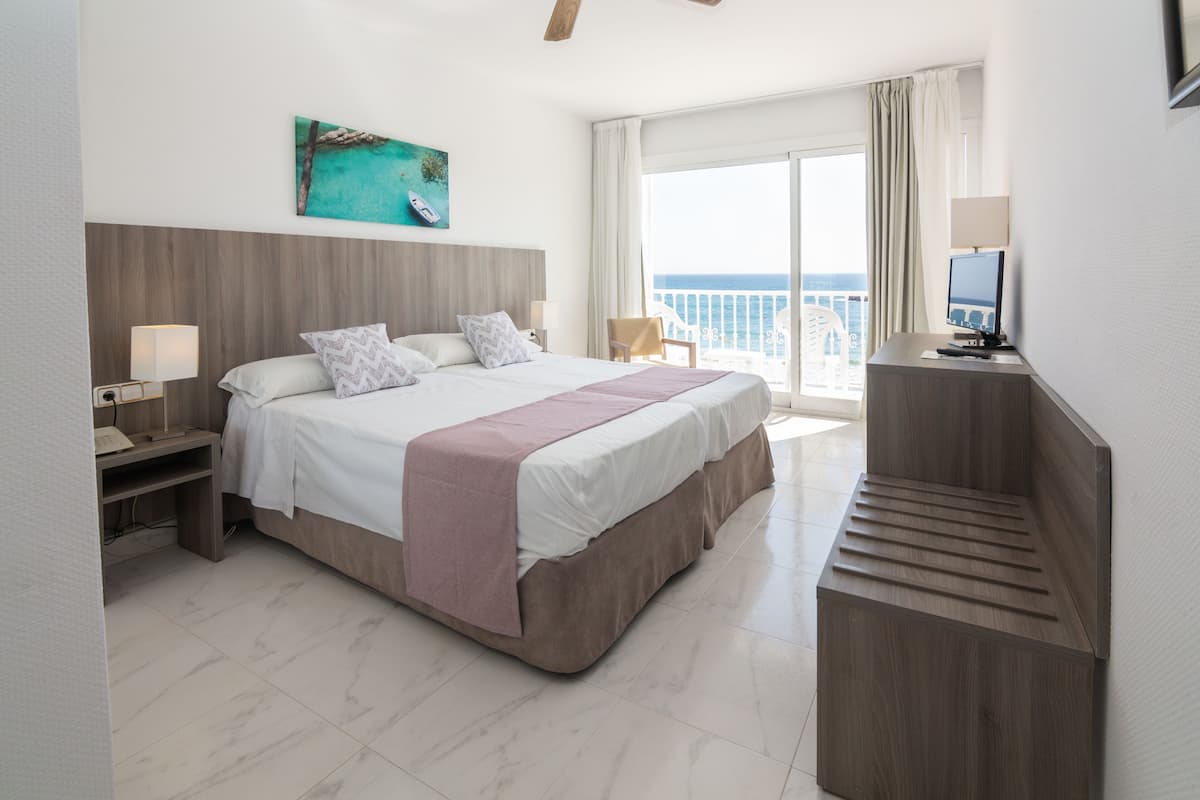Habitación de hotel con una cama grande, un escritorio con ordenador, televisión y balcón con vistas al mar.
