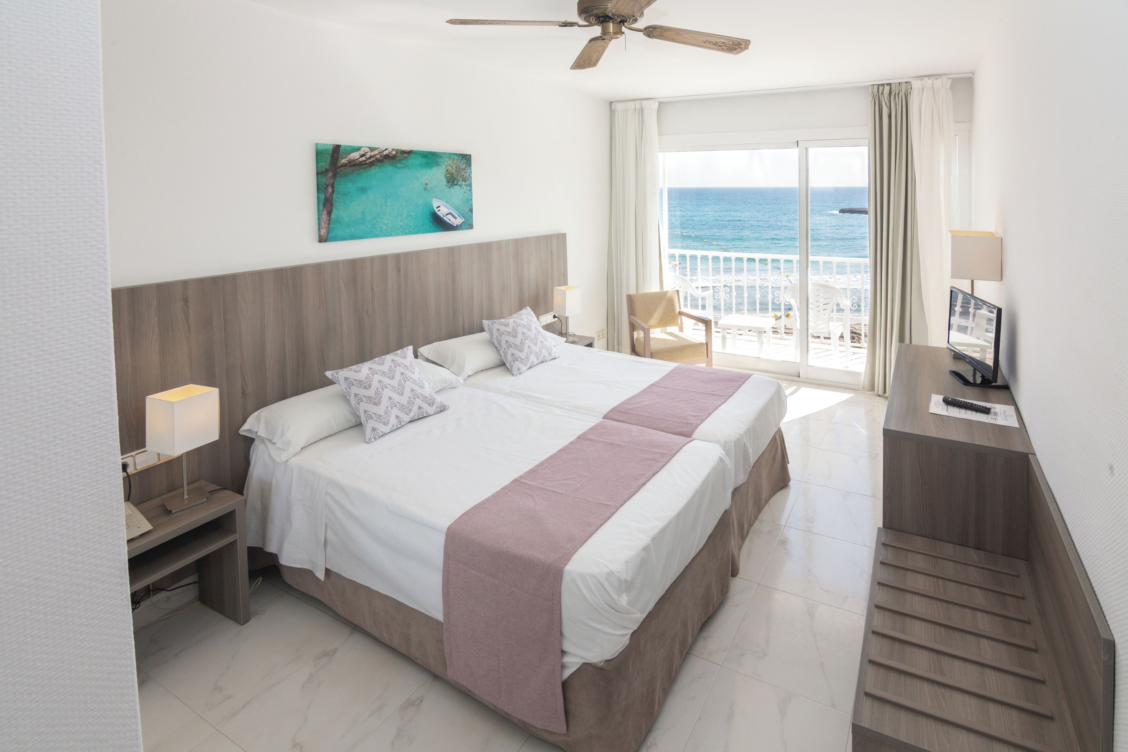 Habitación de hotel con cama, escritorio con computadora, silla, ventana con vista al mar y ventilador de techo.