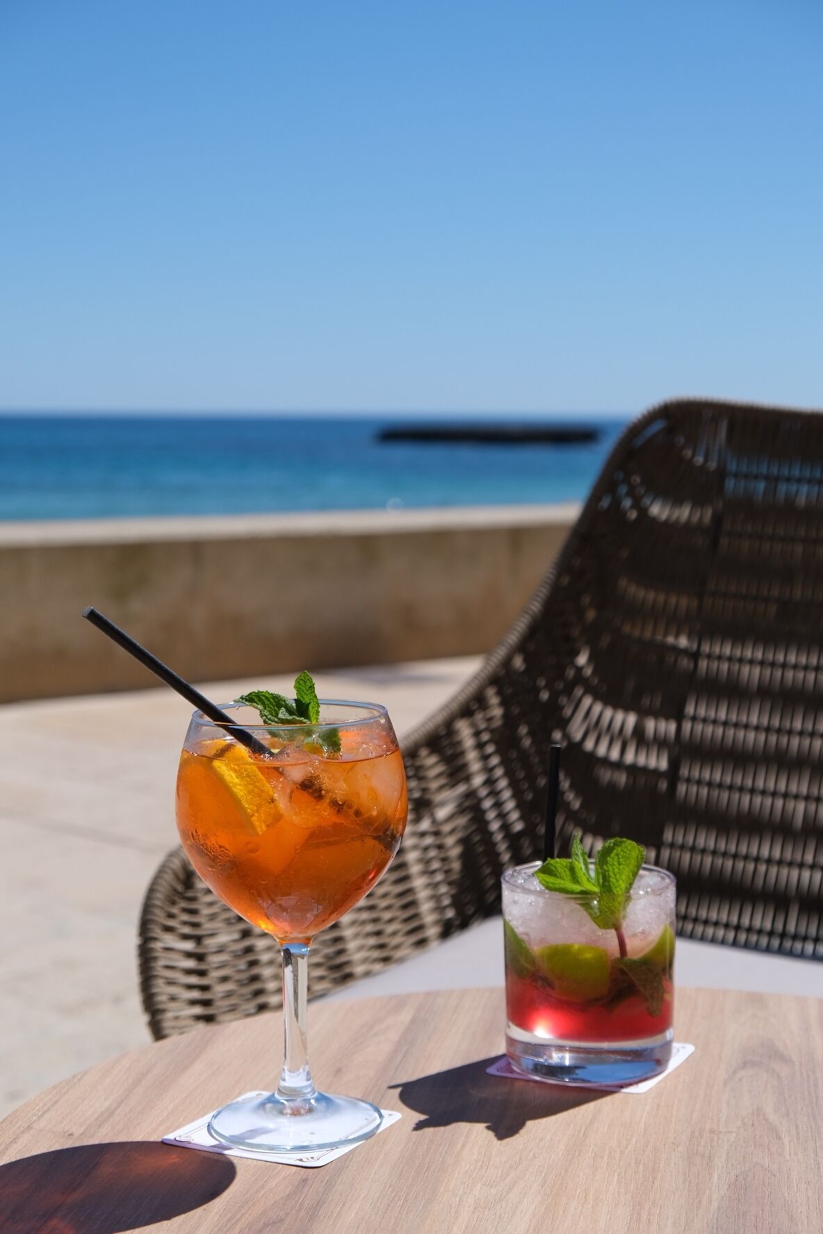 Twee drankjes op een houten tafel, met een rieten stoel op de achtergrond en uitzicht op een strand.