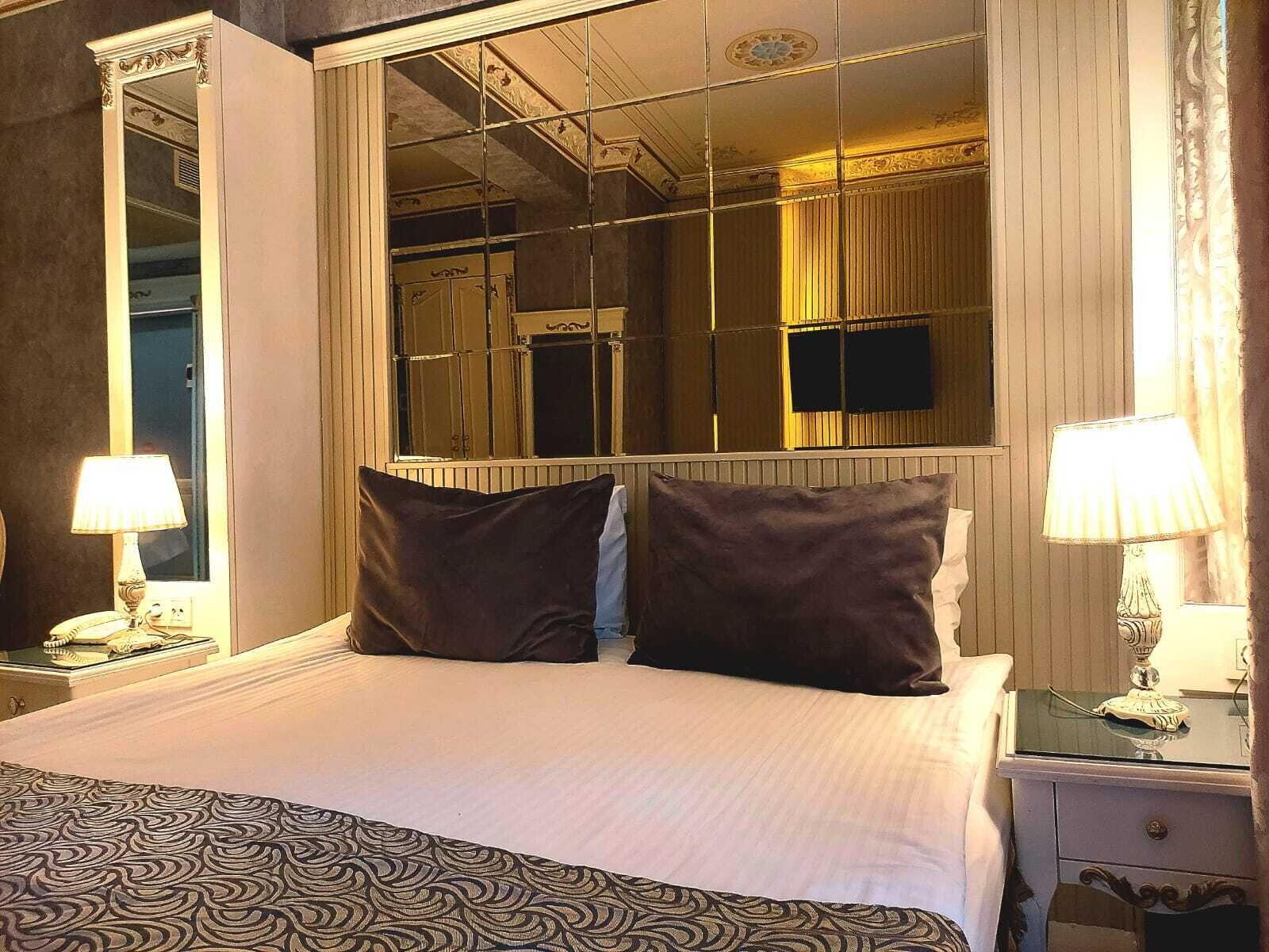 A hotel room with a large mirror, a bed with dark pillows, a patterned throw, and two bedside lamps.