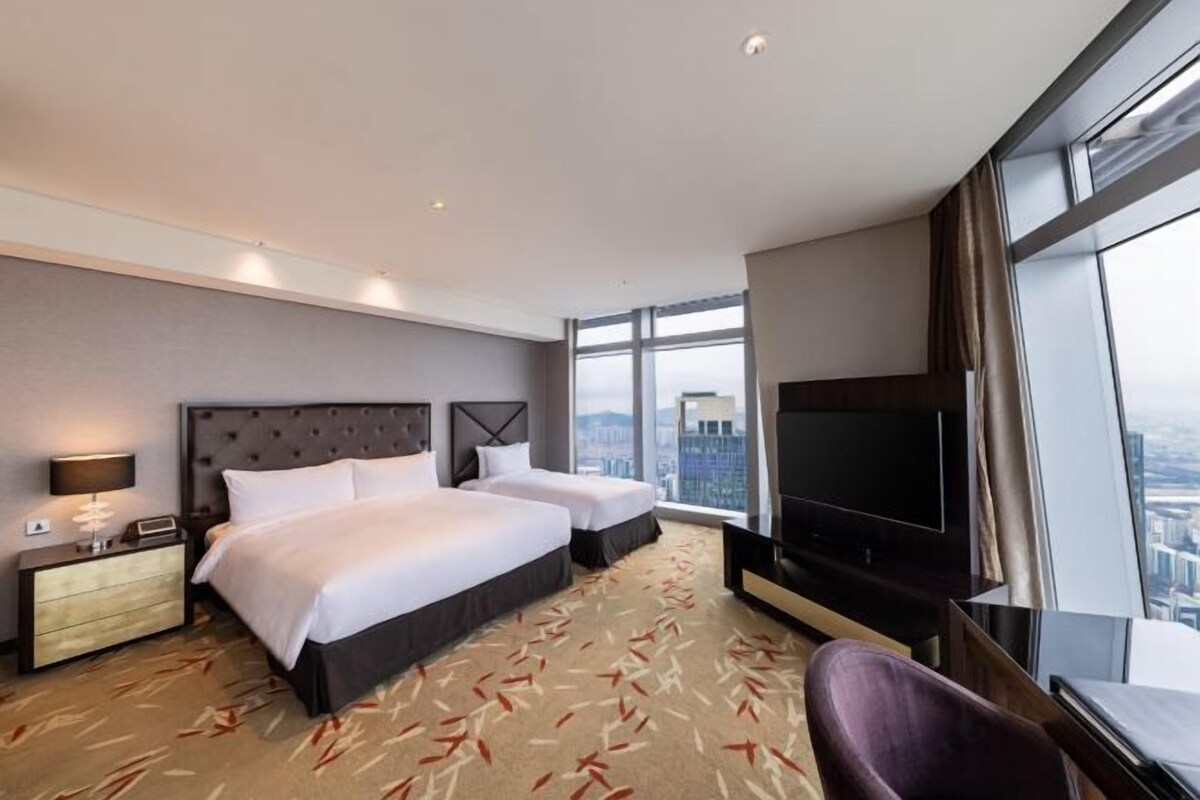 Habitación de hotel con dos camas, un televisor de pantalla plana, un amplio ventanal con vista a la ciudad y piso alfombrado.