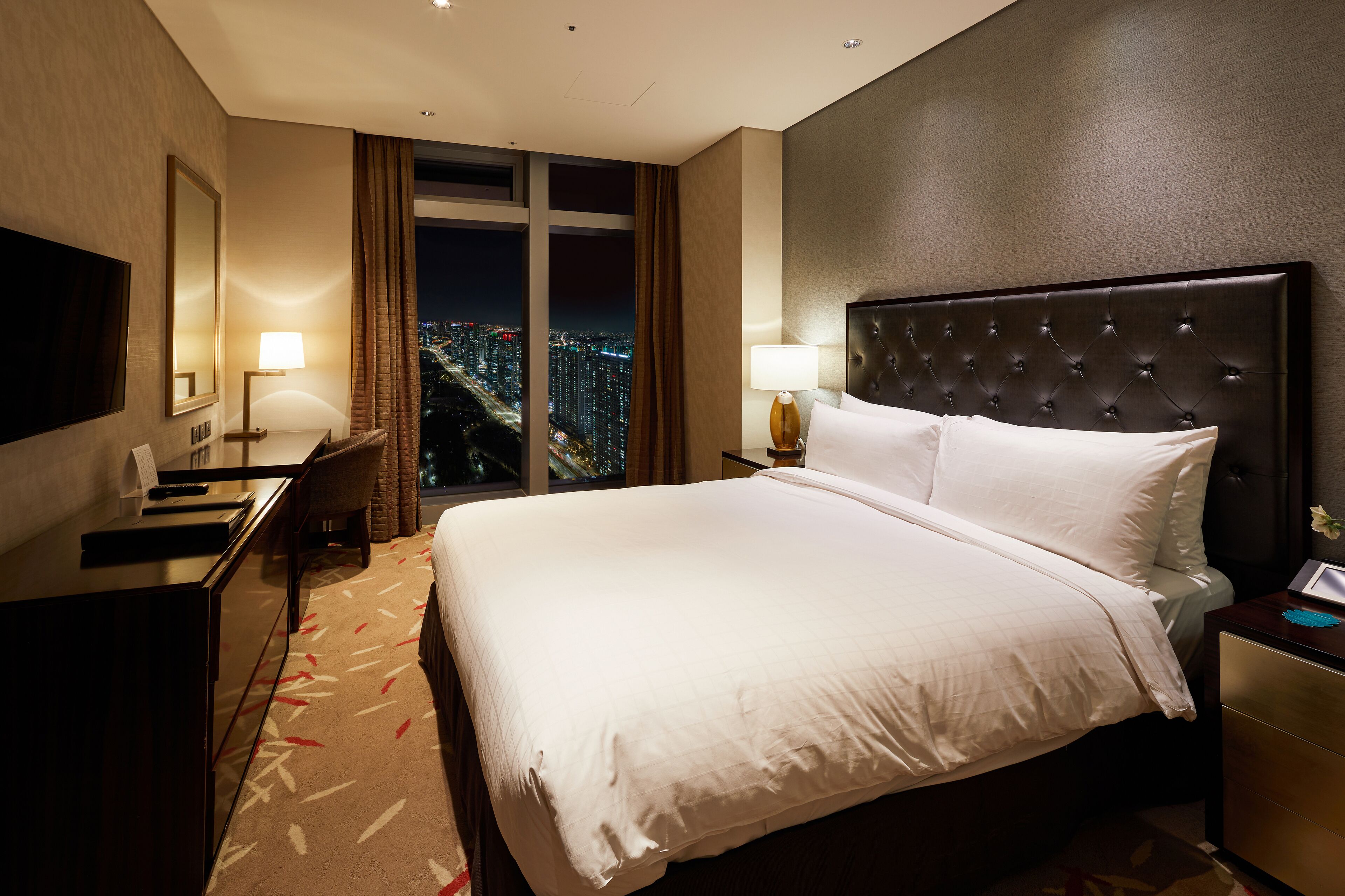 Habitación de hotel con una cama grande, un escritorio, una silla, un televisor y vistas a la ciudad de noche.