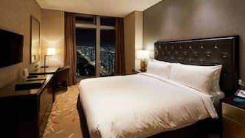 Habitación de hotel con una cama grande, un escritorio, una silla, un televisor y vistas a la ciudad de noche.