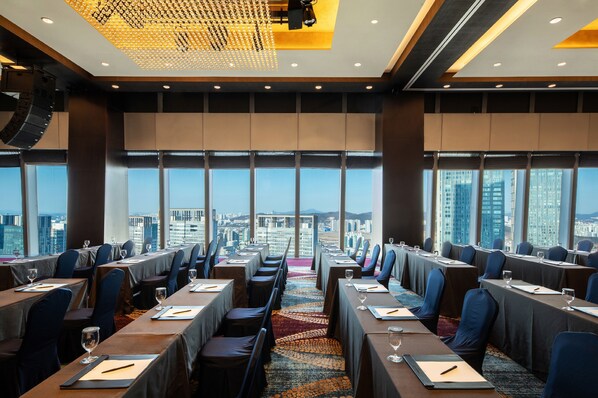 Un salón de conferencias con mesas dispuestas para reuniones, amplias ventanas con vista a la ciudad y una moderna lámpara de araña.