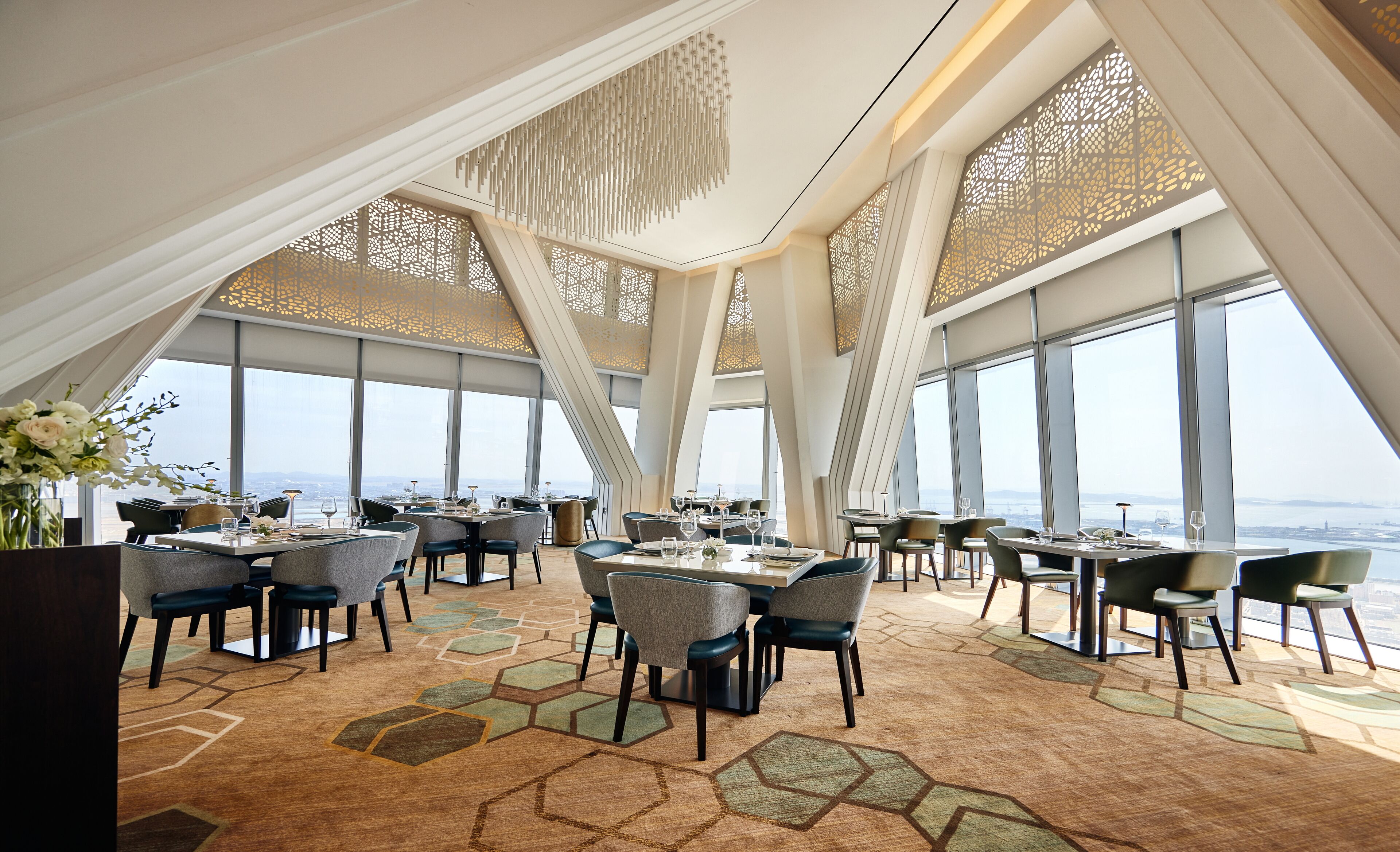 A modern dining area with geometric patterned carpet, high ceilings, and large windows offering a view of the cityscape.