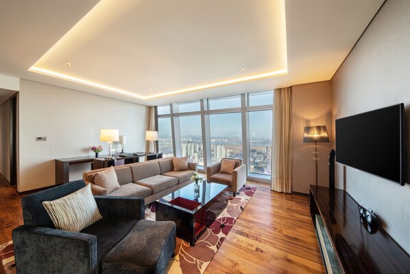 A modern hotel room with a large sofa set, a coffee table, a flat-screen TV, and a city view through large windows.
