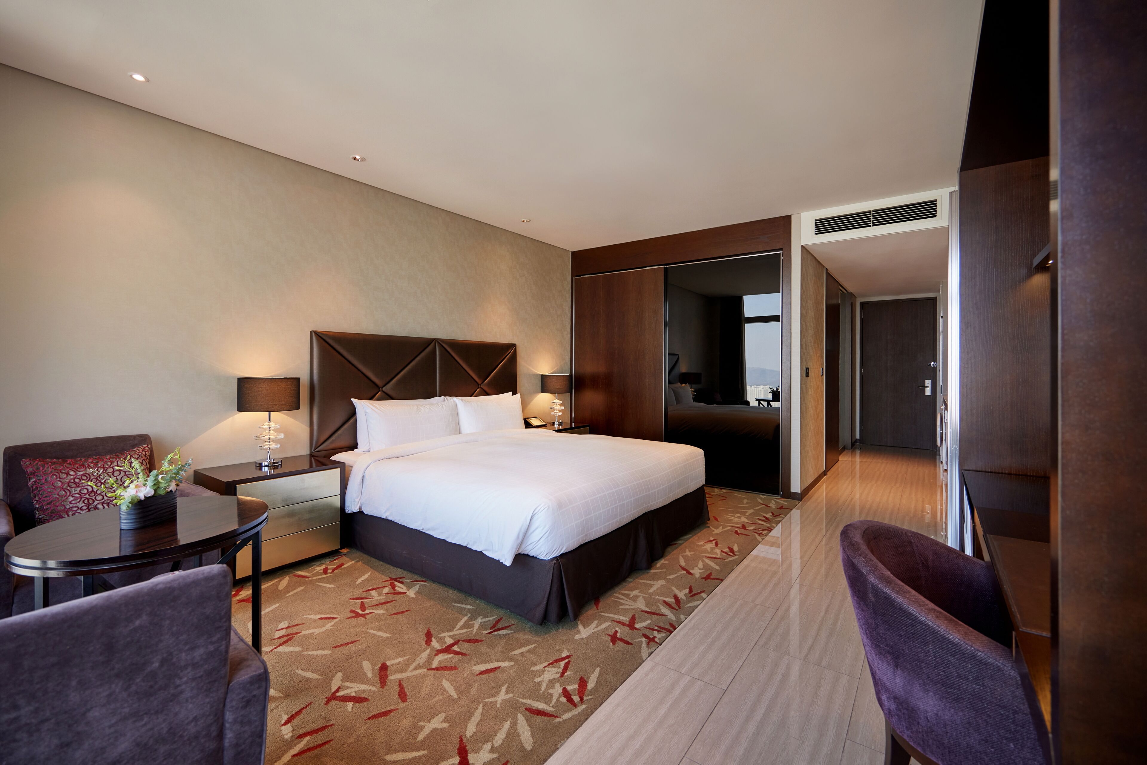 A modern hotel room with a large bed, bedside tables with lamps, a small round table, and a chair.