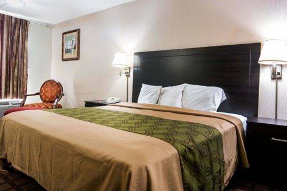 Una habitación de hotel con una cama, dos lámparas de noche, una mesita de noche, una silla y una ventana con cortinas.