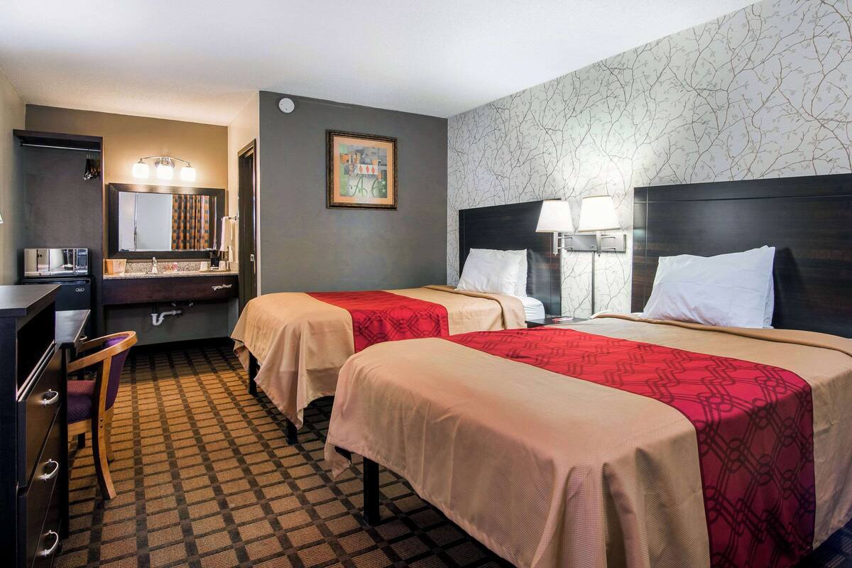 Habitación de hotel con dos camas, un escritorio, una silla, un espejo y una pared con papel tapiz estampado.