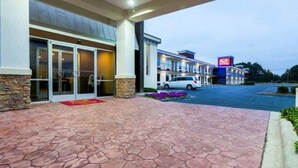 A hotel entrance with a stone-tiled walkway, a car parked in front, and a building with a sign in the background. - Econo Lodge Selma - Smithfield (Selma)