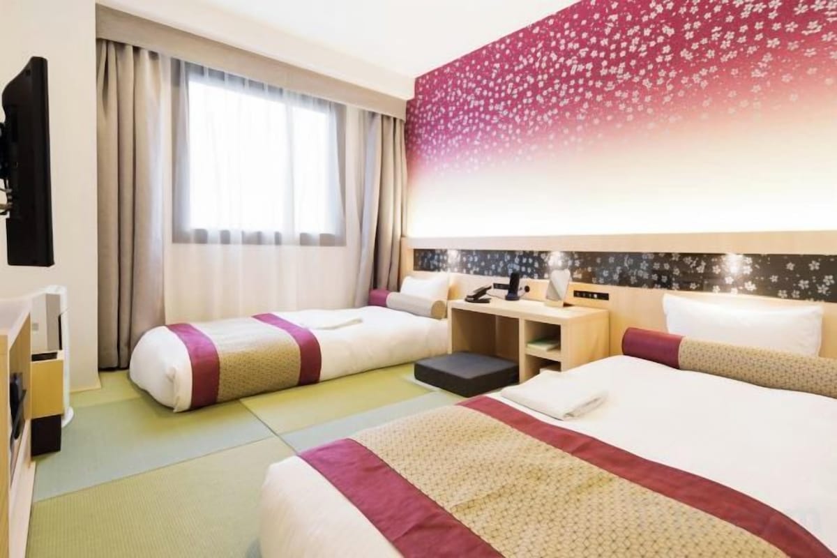 Ein Hotelzimmer mit zwei Betten, einem Schreibtisch, einem Stuhl, einem Fernseher und einem Fenster mit Vorhängen.
