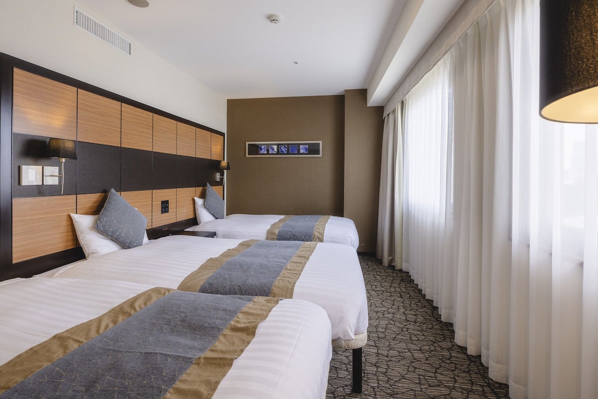 Ein Hotelzimmer mit zwei Betten, einem Kopfteil mit Bedienfeld, einem an der Wand montierten Fernseher und einem Fenster mit Vorhängen.