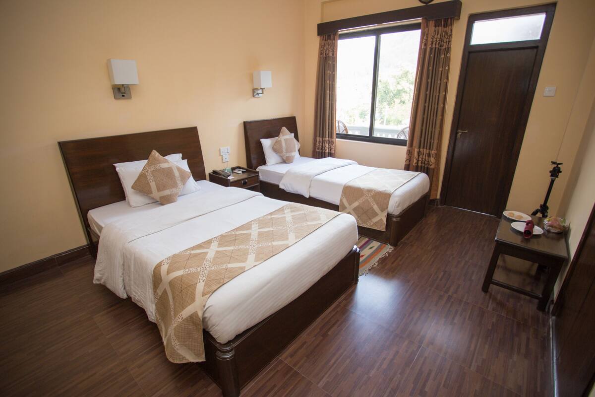 A hotel room with two beds, a window with curtains, a wooden floor, and a small table with a lamp.