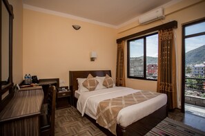 A hotel room with a bed, a desk, a chair, a nightstand, a window with curtains, and an air conditioning unit. - Hotel Peninsula (Pokhara)