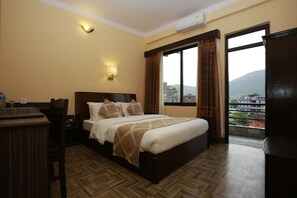 A hotel room with a bed, bedside table, lamp, and a view of the city through a large window. - Hotel Peninsula (Pokhara)