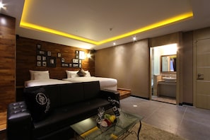 Una habitación de hotel moderna con una cama, un sofá negro, una mesa de centro de vidrio con frutas y una copa de vino, y un baño visible a través de una puerta abierta.