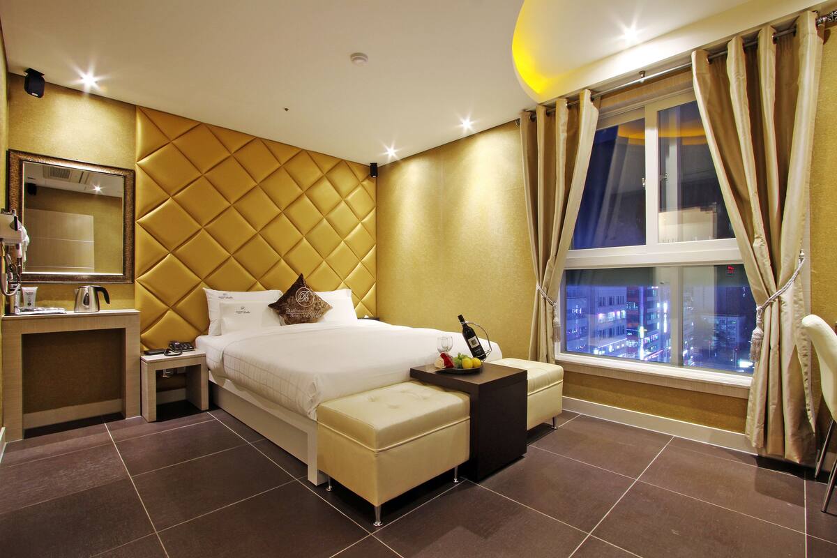 A modern hotel room with a large bed, a bedside table, a small table with a bottle and fruit, and a window with curtains.